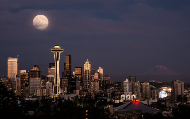 The Space Needle stands tall among city buildings under a bright full moon at night