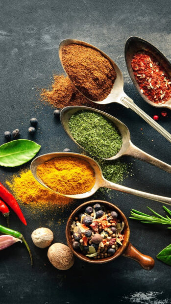 Various colorful spices and herbs arranged in spoons on a dark surface showing vibrant seasoning options