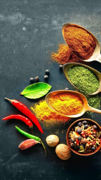 A variety of spices including ground turmeric and chili peppers with fresh herbs and whole seeds on a dark surface