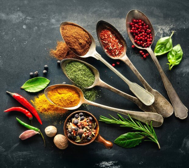 Various spices and herbs in spoons and bowls arranged on a dark surface with fresh chili peppers and leaves