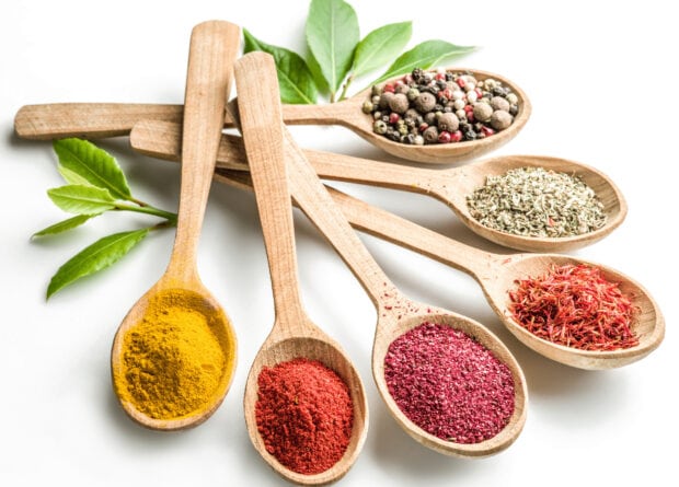 A collection of colorful spices in wooden spoons with fresh green leaves on a white surface