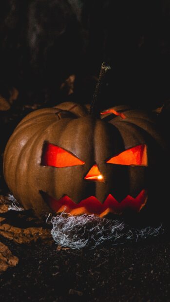 A glowing Halloween pumpkin with a scary carved face sitting in the dark soil, 4K Phone Wallpaper