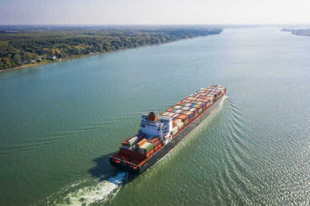 A large cargo ship loaded with containers sails on the St Lawrence River near a forested shoreline