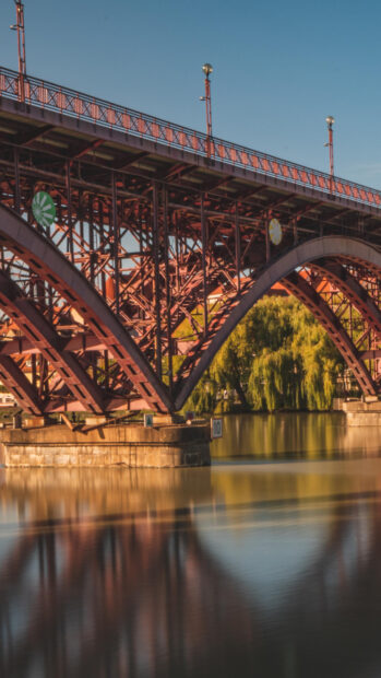 A close up view of a red metal bridge over the calm St Lawrence River with reflections and trees in the background