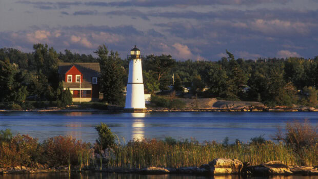 A white lighthouse and red house stand by the St Lawrence River under a partly cloudy sky at sunset