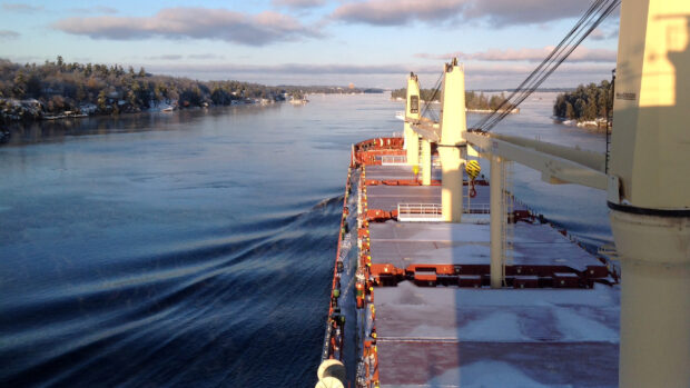 A large cargo ship sails down the calm St Lawrence River surrounded by forested shores under a clear sky