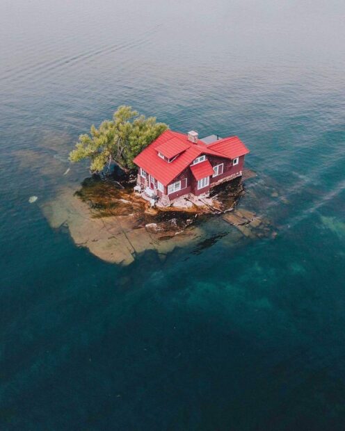 A small red roofed house sits on a rocky island surrounded by the clear waters of the St Lawrence River