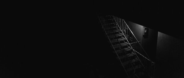 A dark metal staircase with railings illuminated by a single light fixture in low light conditions