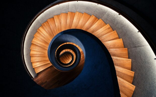 A spiral staircase made of wood winding downward with a blue central structure and ambient lighting around the steps