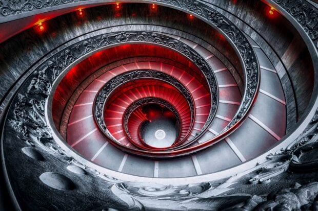 A spiral staircase with detailed stone carvings illuminated by red lights viewed from above