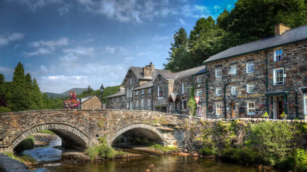 A stone bridge crosses a river near old buildings in a scenic Wales village with green trees, HD Desktop Wallpaper