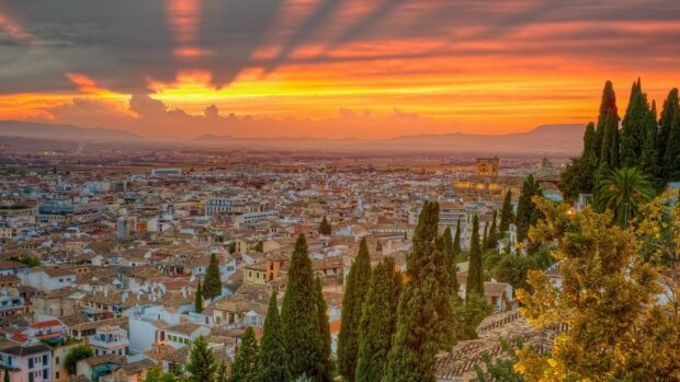 A breathtaking view of a Spanish city under a vibrant sunset sky with lush green trees, HD Desktop Wallpaper