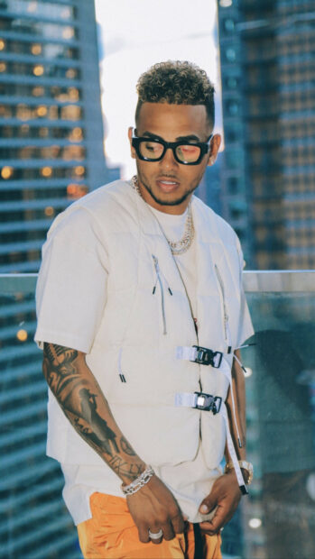 A stylish man wearing glasses and chains stands with Ozuna Wallpapers in front of tall buildings, HD Mobile Wallpaper