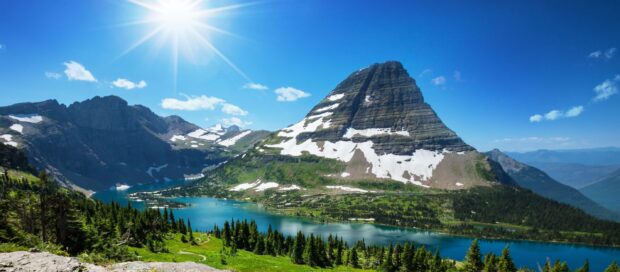 A bright sunny day shows the mountain and lake at Glacier National Park with green trees and blue sky, 2K Desktop Wallpaper