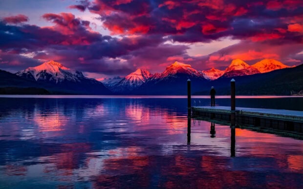A person stands on a dock by a lake reflecting pink and purple skies with Montana landscapes, HD Desktop Wallpaper