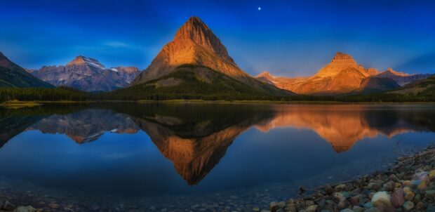 The sun sets behind towering peaks with a clear blue sky at Glacier National Park, HD Desktop Wallpaper