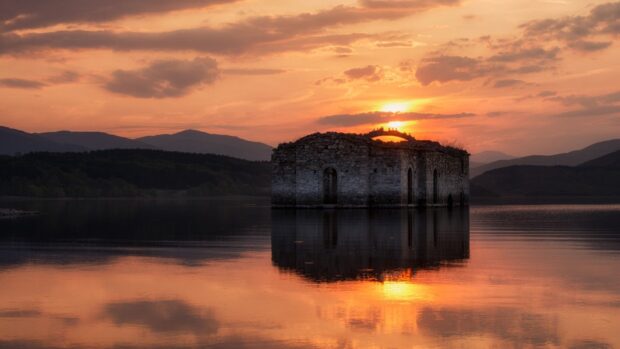 A calm water lake surrounds an old stone building during a vivid sunset in Bulgaria, HD Desktop Wallpaper