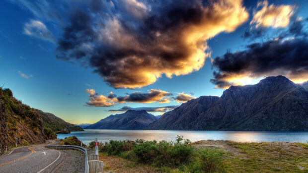 A winding mountain road runs alongside a peaceful lake beneath dramatic sunset clouds with mountain background, 4K Desktop Wallpaper