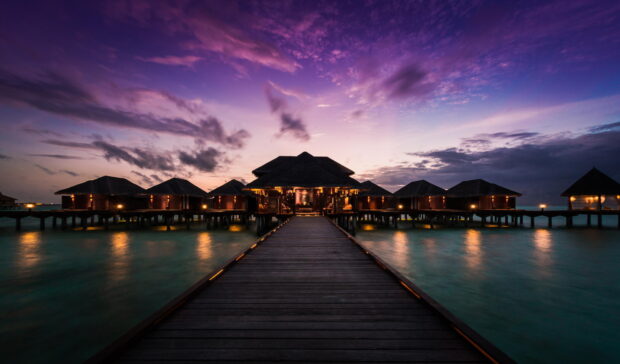 A peaceful bungalow is shown at sunset with glowing lights over calm water, HD Desktop Wallpaper