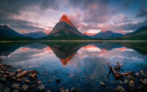 A mountain peak glows under the sunset sky reflecting in clear lake water at Glacier National Park, 2K Desktop Wallpaper