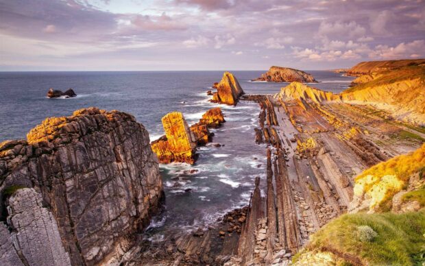 A rocky coastline in Spain glows under a purple sunset sky with calm ocean water, 2K Desktop Wallpaper