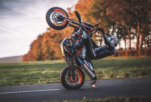 A supermoto rider performing a wheelie on a road with autumn trees in the background