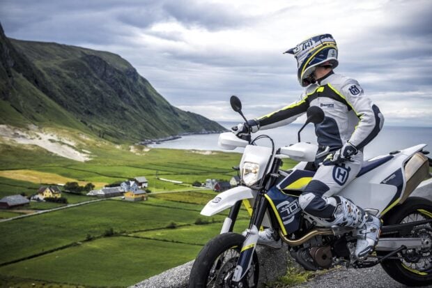 Rider on supermoto motorcycle overlooking green valley and sea with mountains in background