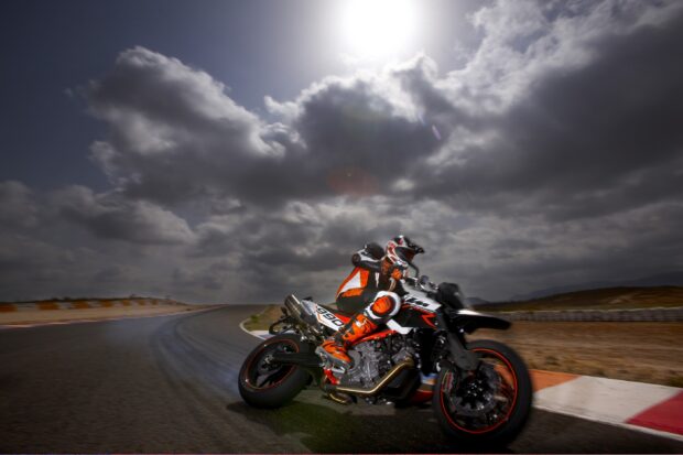 A supermoto rider wearing orange gear leans into a sharp turn on the racetrack under a cloudy sky