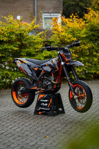 Orange and black supermoto bike on a stand in outdoor setting with autumn leaves background