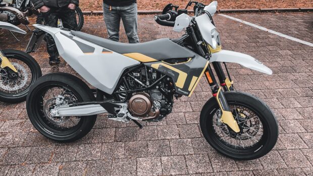 A supermoto motorcycle with yellow and gray details parked on a brick pavement with two people standing nearby