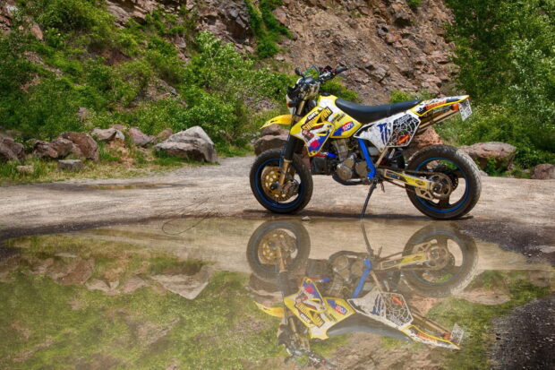 A supermoto bike parked on a dirt road with its reflection in the water