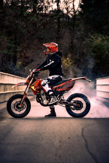 Rider wearing an orange helmet performing a stunt on a supermoto bike on the bridge