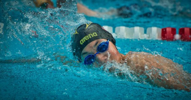 A swimmer wearing blue goggles and a swimming cap swimming freestyle in a pool with clear water