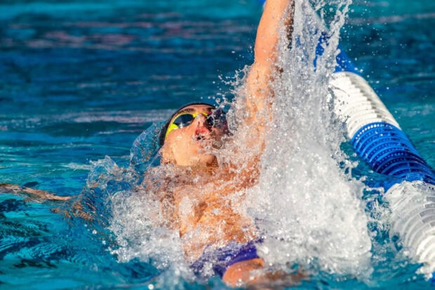 A swimmer wearing goggles performs backstroke swimming in a clear pool with splashing water