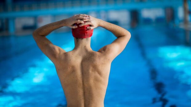 Male swimmer wearing a red swim cap standing at the poolside with hands behind head