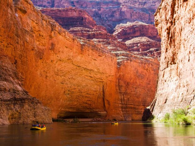The Colorado River flowing through the canyon with towering red rock walls and people rafting on the water