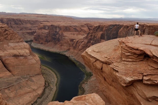 A vast desert canyon with the Colorado River winding through and a person standing on the cliff edge overlooking the landscape