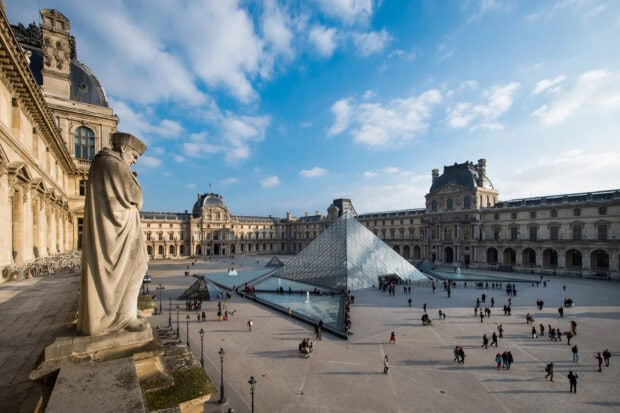 Statue overlooking the Louvre courtyard with glass pyramid under a blue sky