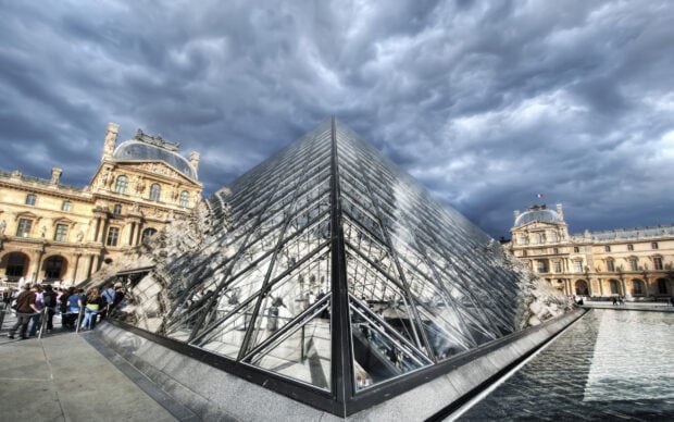 The Louvre glass pyramid under a cloudy sky with Paris museum buildings and visitors