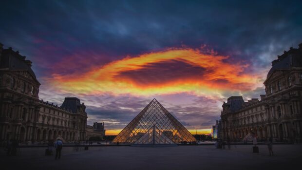 The Louvre glass pyramid illuminated at sunset with dramatic sky clouds and historic buildings