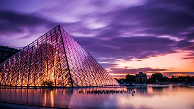 The Louvre glass pyramid illuminated at sunset with reflective water and dramatic clouds