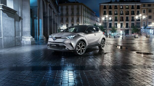 Compact silver Toyota C-HR parked on wet cobblestone street at night in urban cityscape with tall buildings and streetlights.