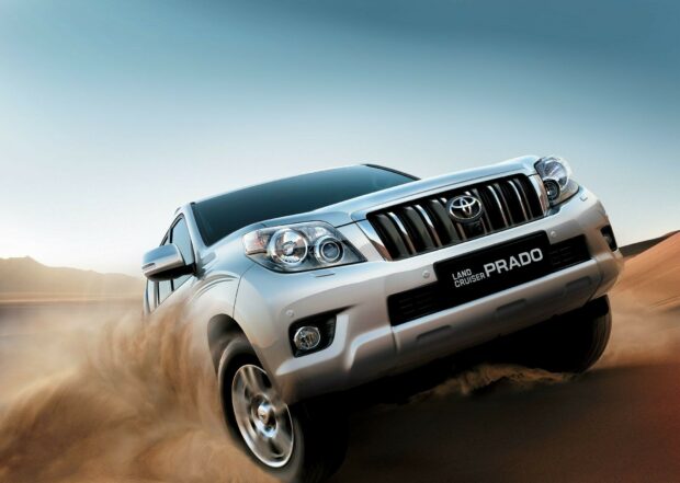 Toyota Land Cruiser Prado SUV driving on sandy desert terrain, showcasing off-road capability and rugged design, with a clear sky background.