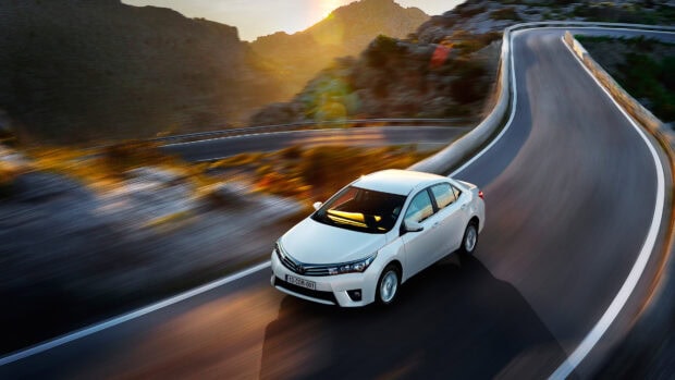 Compact white sedan driving on a winding mountain road at sunset with scenic landscape and guardrails in the background.