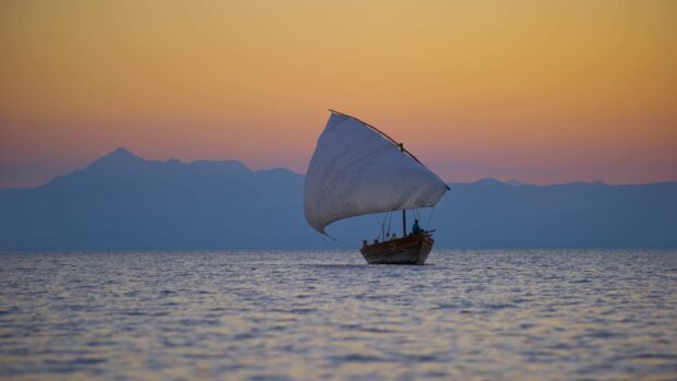 A traditional sailboat is sailing on the calm waters with mountains in the background at sunset, HD Desktop Wallpaper