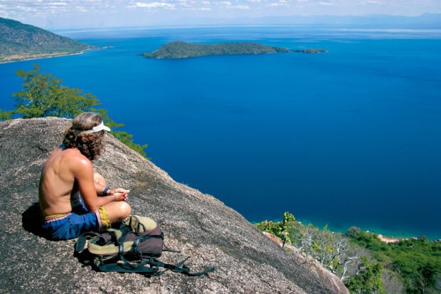 A man sits on a rocky hill overlooking the deep blue lake in Malawi Wallpapers, 2K Desktop Wallpaper