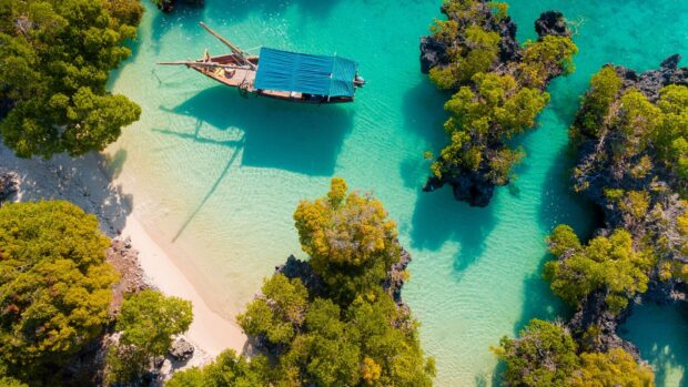 A traditional wooden boat sails through clear turquoise waters surrounded by lush greenery in Zanzibar, HD Desktop Wallpaper