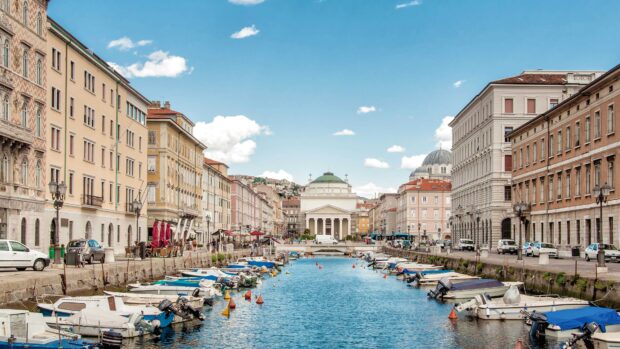 A view of Trieste with boats lined along a canal and historic buildings under a blue sky, 4K Desktop Wallpaper