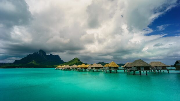 The bungalow is located over the turquoise water with a mountain in the background under a cloudy sky