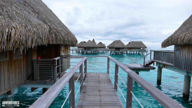 A wooden pathway leads to bungalow houses over clear turquoise water, 4K Desktop Wallpaper